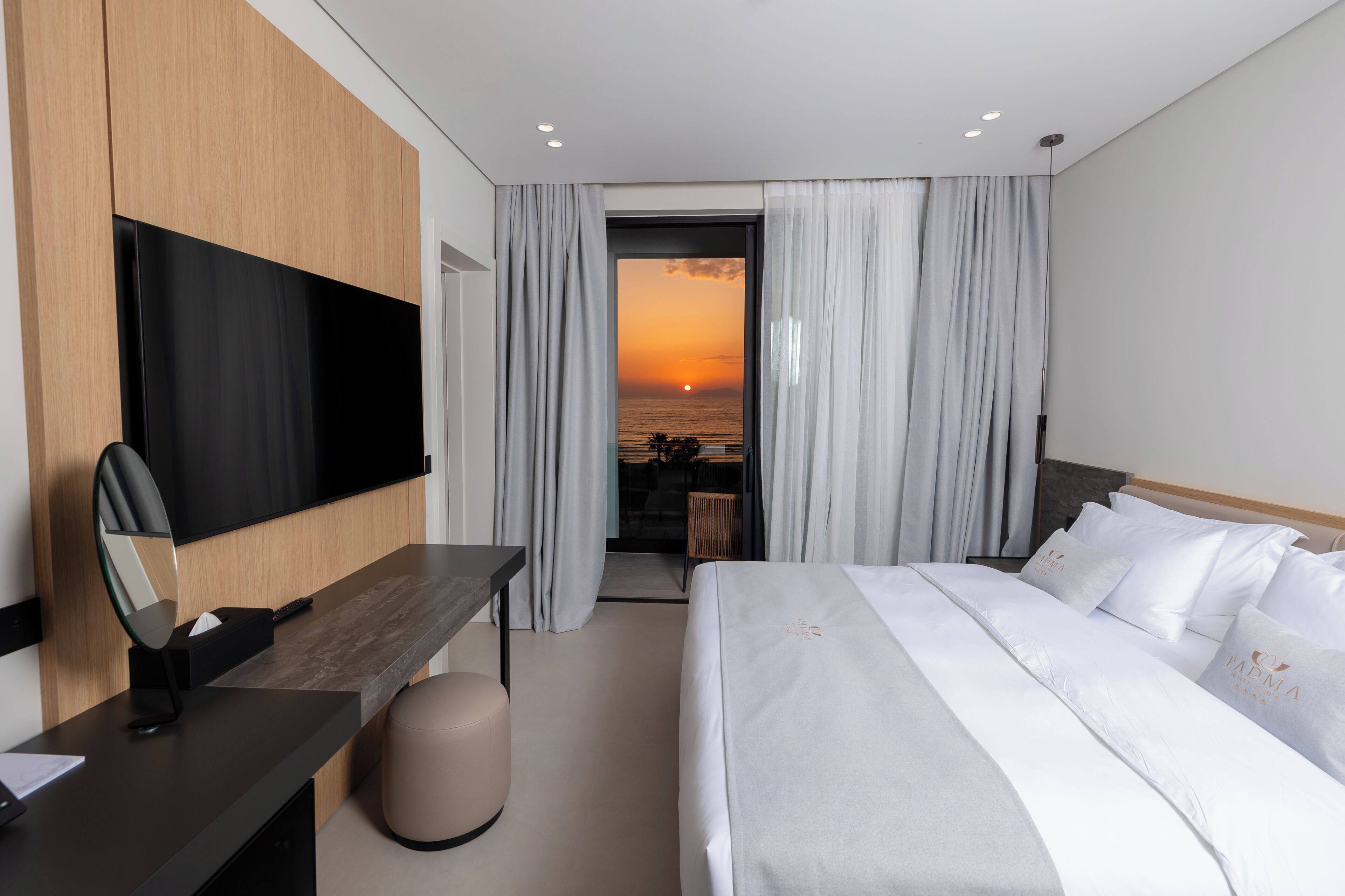 Een moderne hotelkamer met een bed, een bureau, een televisie en uitzicht op de ondergaande zon boven de oceaan.