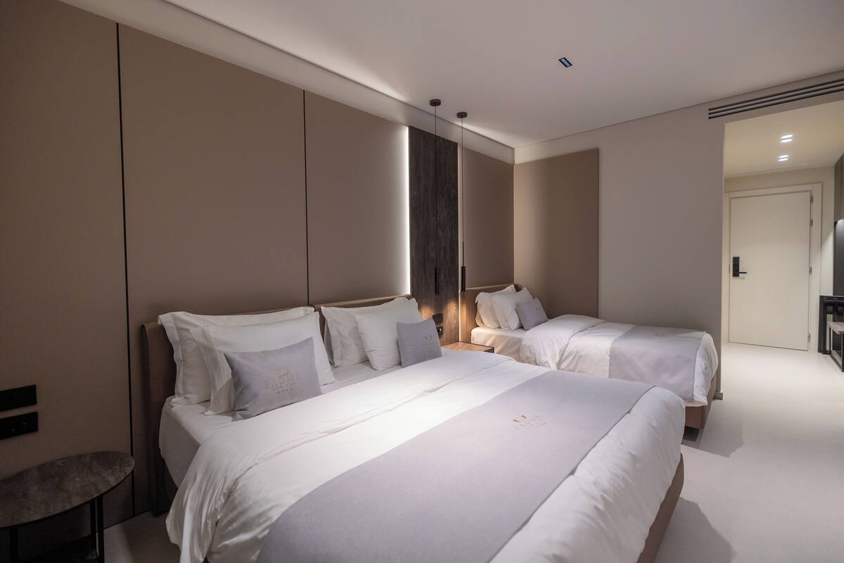 A modern hotel room with two beds, a bedside table, a chair, and a large mirror.