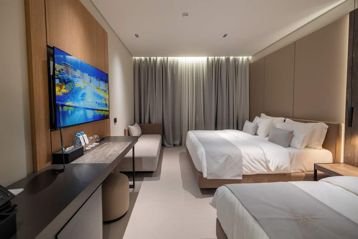 A modern hotel room with a flat-screen TV, a desk with a chair, two beds, and a large window with curtains.