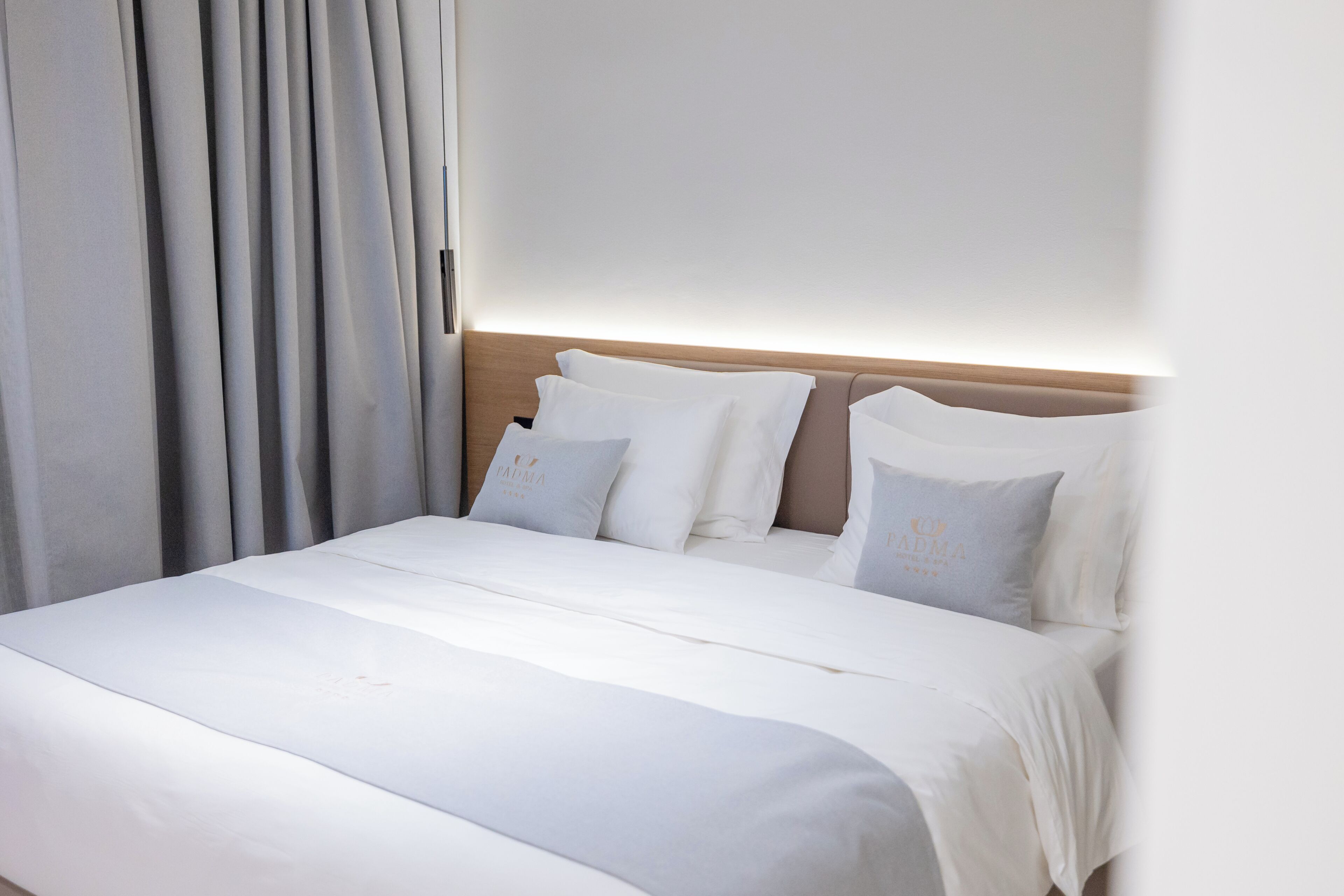 A neatly made bed with white linens and pillows, a wooden headboard, and a large window with curtains.