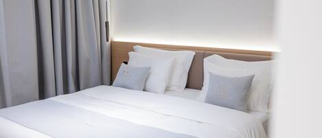Una cama bien hecha con sábanas y almohadas blancas, cabecera de madera y un amplio ventanal con cortinas.