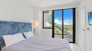 Une chambre moderne avec un grand lit, une tête de lit bleue et une porte-fenêtre coulissante donnant sur un balcon avec vue sur la ville.