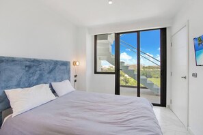 A modern bedroom with a large bed, a blue headboard, and a sliding glass door leading to a balcony with a city view. - Penthouse Duplex w/ Large Terrace, Pool, Golf and Ocean Views (Punta Cana)