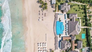 Vue aérienne d’un complexe hôtelier de bord de mer, avec une piscine, des chaises longues et des palmiers.