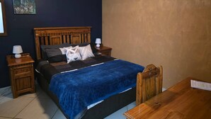 A bedroom with a wooden bed, two nightstands, a blue blanket, and a wooden chair. - The Pot Faerie Glen in Pretoria (Pretoria)