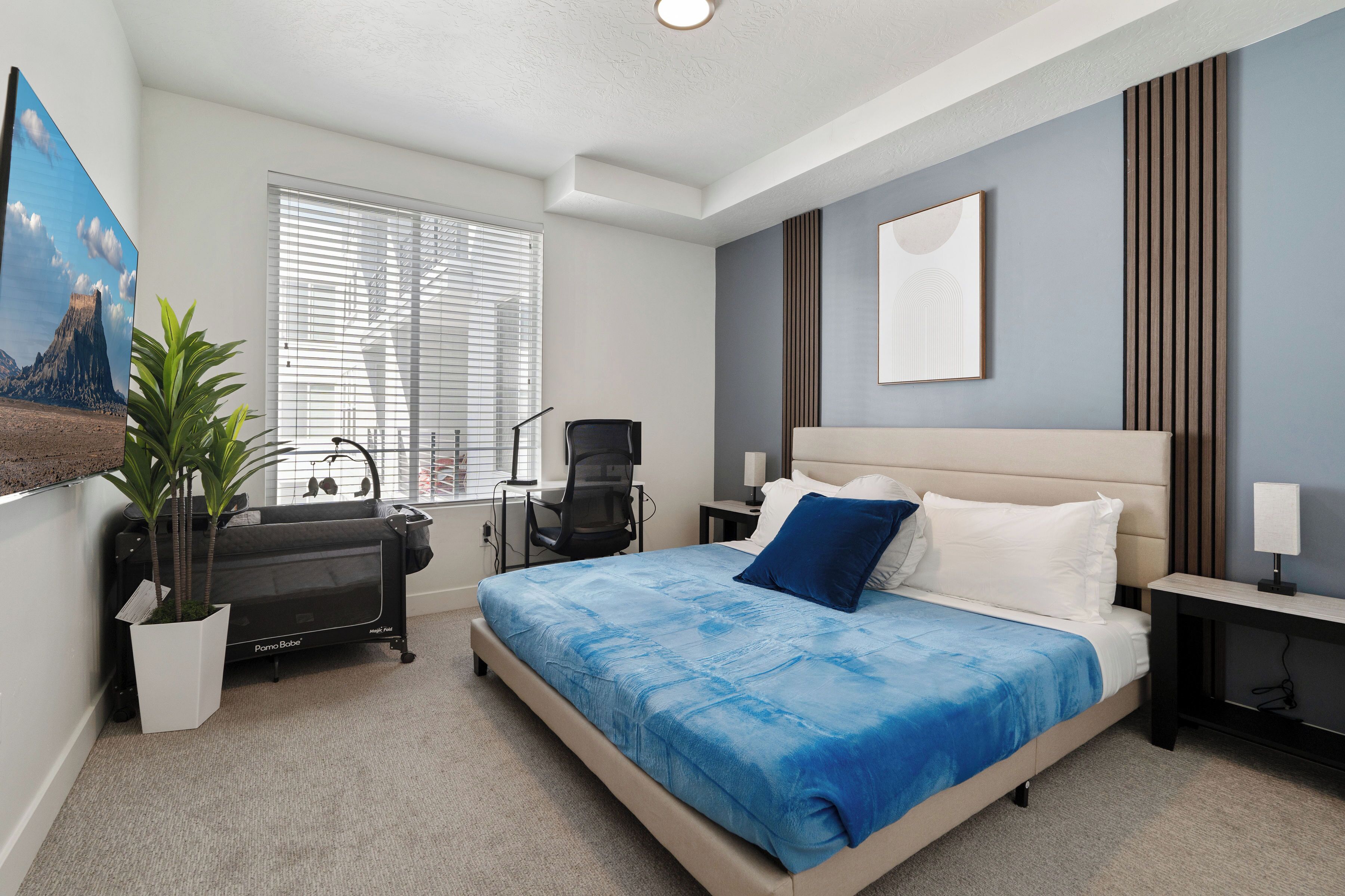 A bedroom with a bed, a desk, a chair, a potted plant, and a window with blinds.