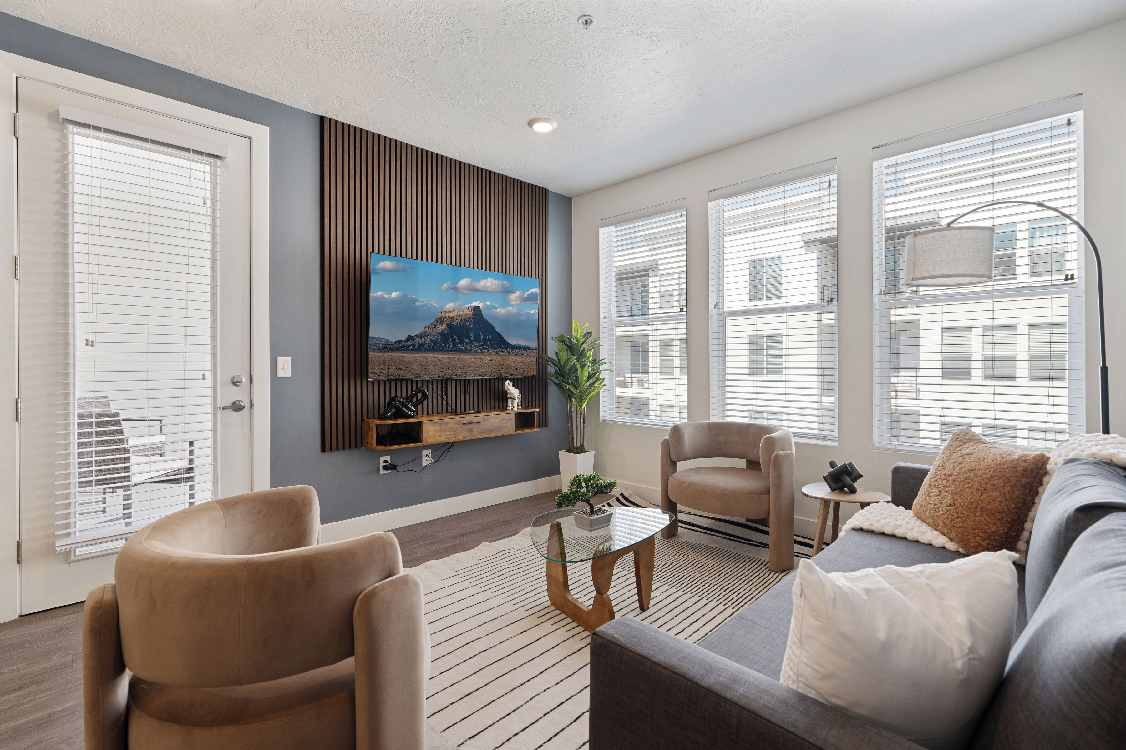 A living room with a sofa, chairs, a coffee table, a TV mounted on the wall, and a window with blinds.