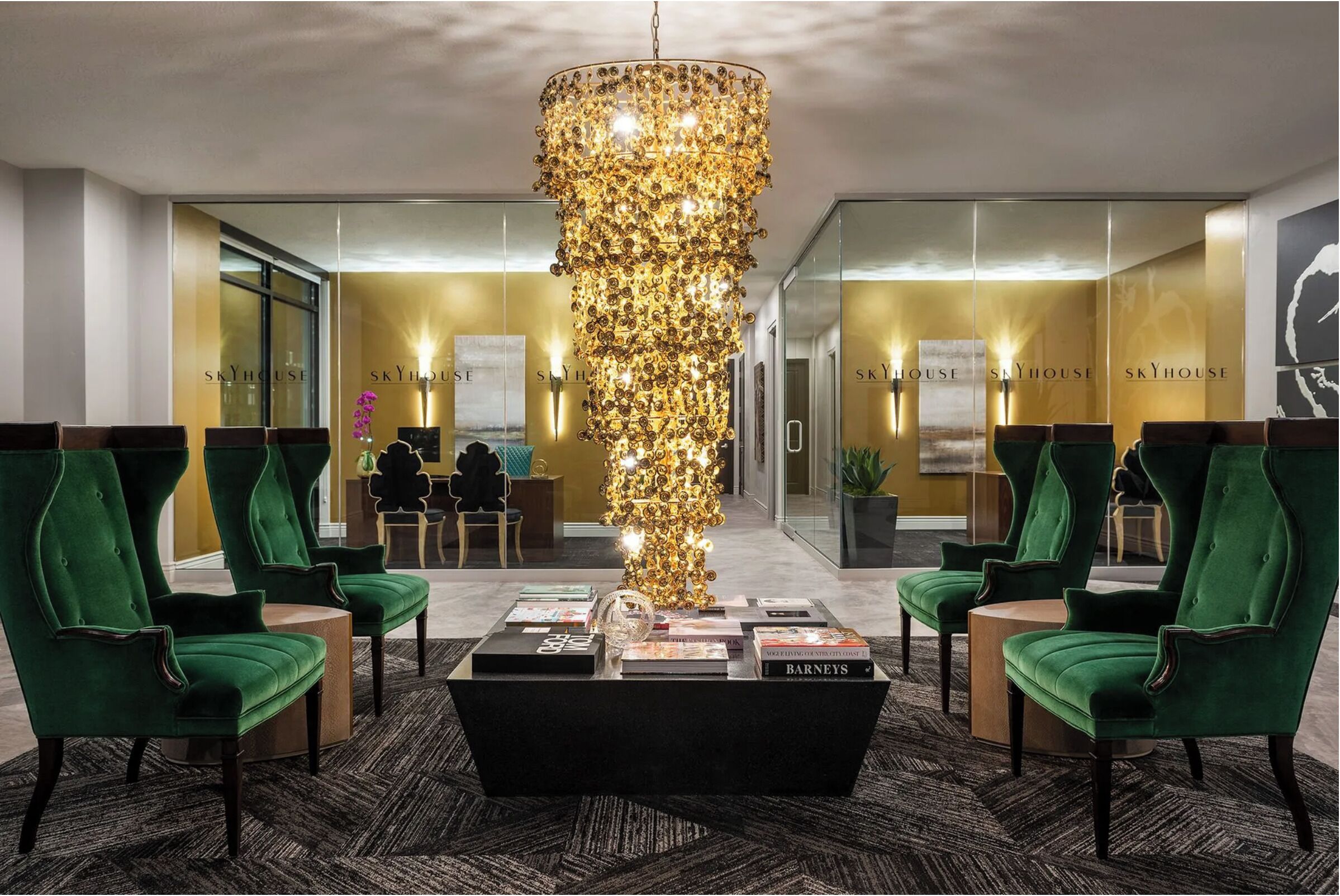 Eine Lobby mit einem großen Kronleuchter, grünen Polsterstühlen, einem schwarzen Couchtisch mit Büchern und einer Glaswand mit der Beschriftung 'SKYHOUSE'.