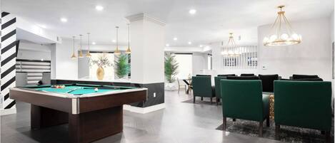 A modern indoor space with a pool table, green chairs, and a dining area with a chandelier.