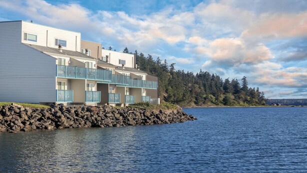 Un immeuble résidentiel situé au bord de l’eau, doté de nombreux balcons, avec une côte rocheuse et une forêt en arrière-plan.