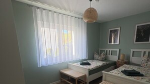 Quarto com duas camas, uma janela com cortinas leves, luminária de fibra natural e tapete felpudo no chão.