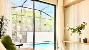 A modern living room with a glass door leading to a pool area, a sofa, a potted plant, and a shelf with decorative items. - Modern Getaway w/Game Room + Pvt Pool + BBQ Near Universal and I-Drive (Orlando)