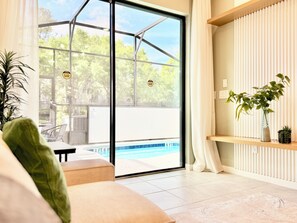 Ein modernes Wohnzimmer mit Glastür zum Poolbereich, einer Couch, einer Topfpflanze und einem Regal mit Dekoration.