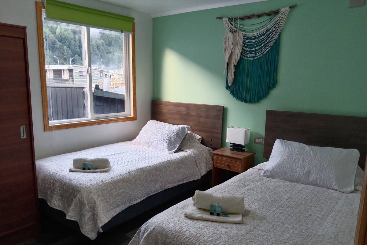 Habitación con dos camas, una ventana, cabecera de madera, mesita de noche y un colgador de macramé en la pared.