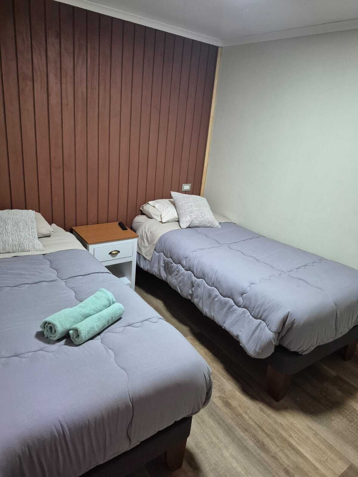 Una habitación con dos camas individuales, una mesita de noche de madera, un cajón blanco y suelo de madera.
