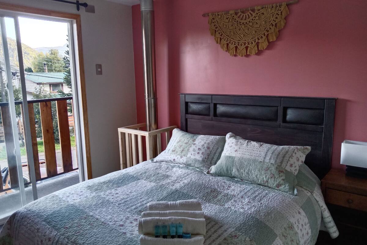 Un dormitorio con cama, almohadas, cabecera, mesita de noche, un adorno en la pared y ventana con vistas.