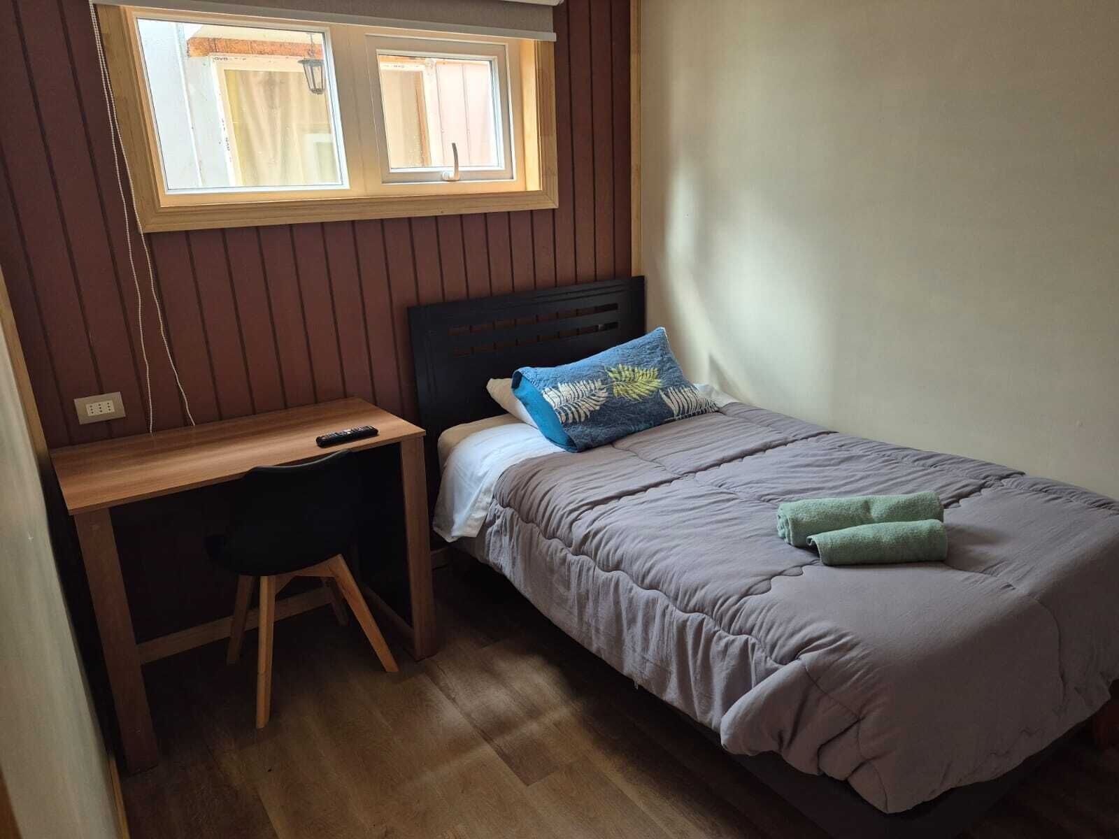 A bedroom with a bed, a desk, a chair, a window, and a wall socket.