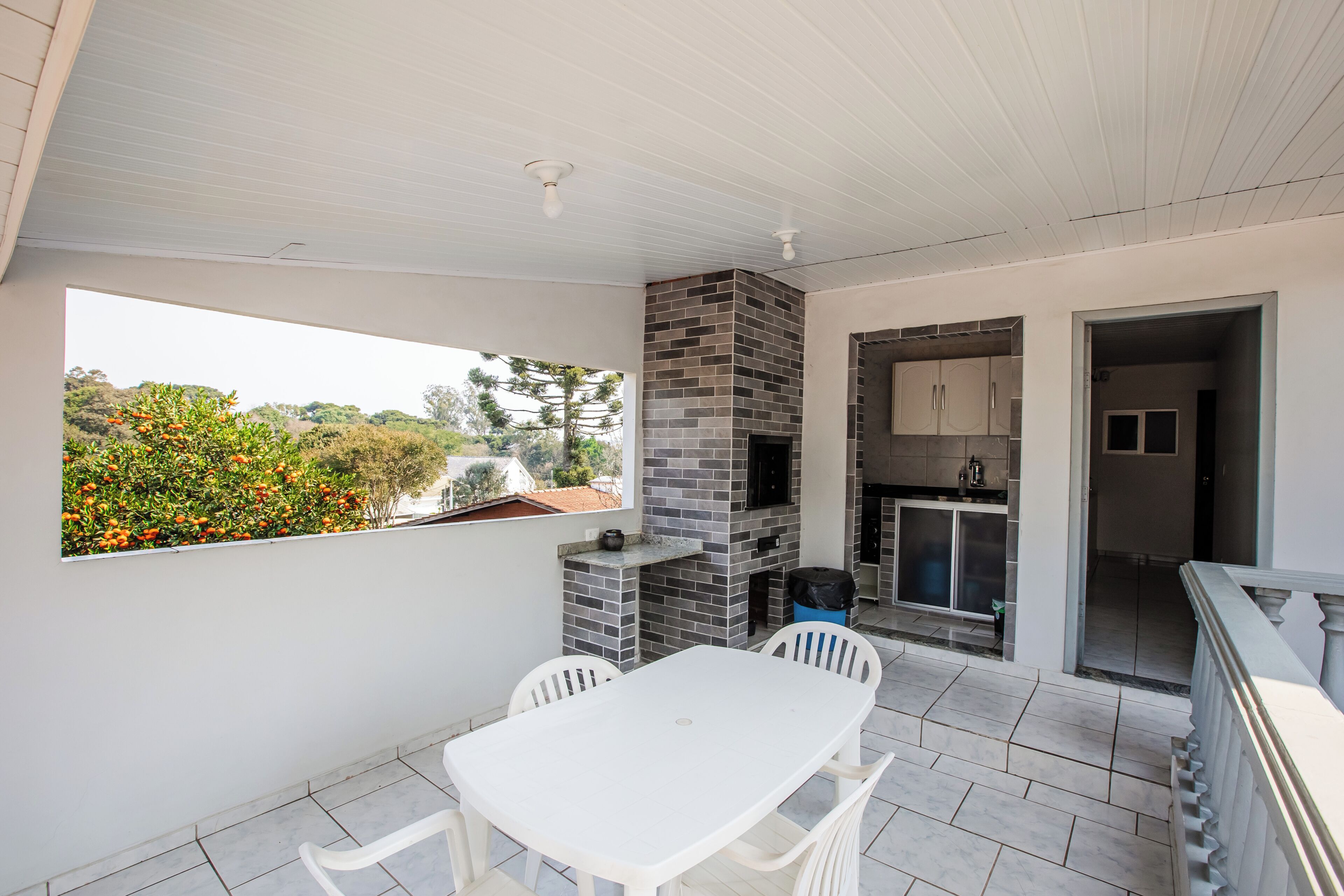 Un patio techado con una mesa y sillas, una chimenea de ladrillo y vistas a árboles y casas.