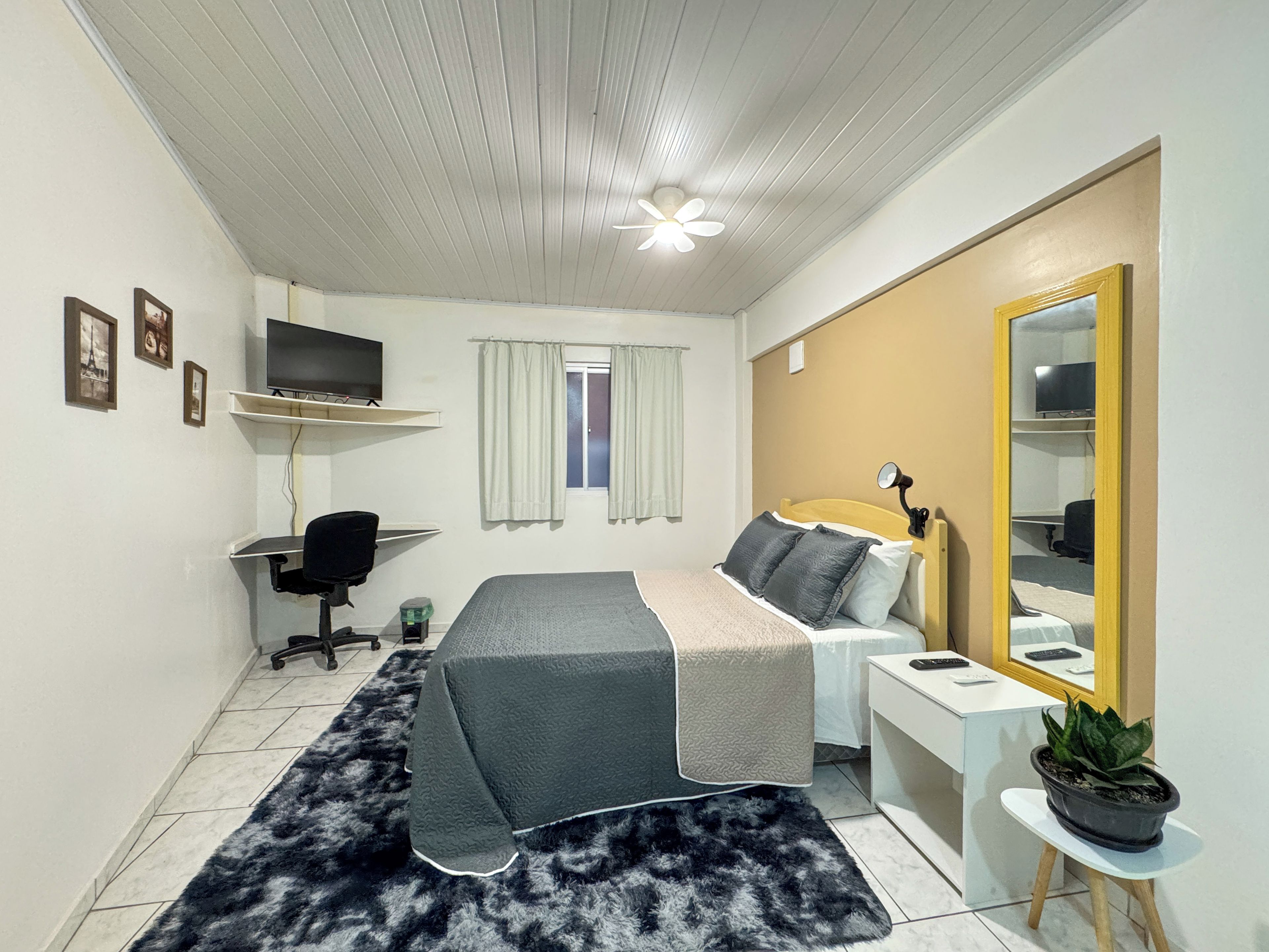 A bedroom with a bed, a desk, a chair, a television, a window with curtains, a ceiling fan, and a potted plant.