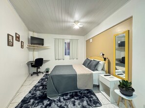 A bedroom with a bed, a desk, a chair, a television, a window with curtains, a ceiling fan, and a potted plant.