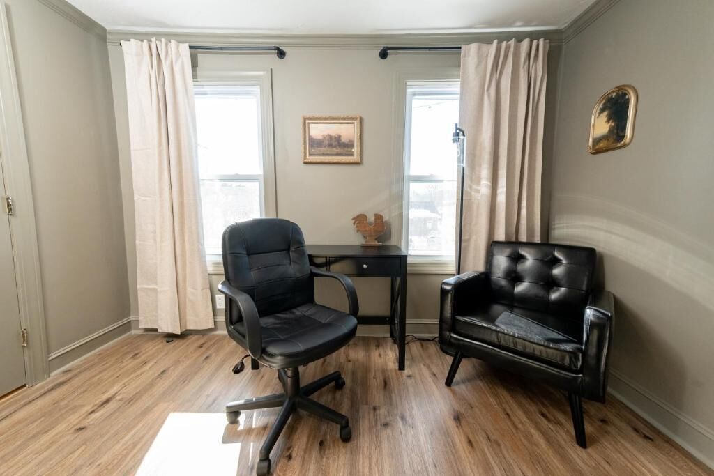 Ein Zimmer mit Schreibtisch, zwei schwarzen Stühlen, einem Fenster mit Vorhängen, einem gerahmten Bild und einem kleinen dekorativen Gegenstand an der Wand.