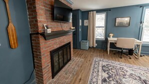 Una habitación con chimenea de ladrillo, televisor montado, escritorio con silla y una alfombra grande.