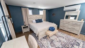 A bedroom with a bed, bedside table, bench, dresser, and an air conditioning unit.