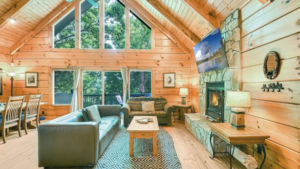 A log cabin interior with a high ceiling, large windows, a fireplace, and a cozy seating area. - Lake Lure Lodge in the Trees Sleeps Eight (Lake Lure)