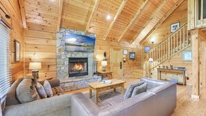 A log cabin interior with a stone fireplace, wooden walls, a sofa, a coffee table, and a staircase. - Lake Lure Lodge in the Trees Sleeps Eight (Lake Lure)