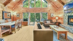 A log cabin interior with a dining area, sofa, fireplace, and large windows. - Lake Lure Lodge in the Trees Sleeps Eight (Lake Lure)