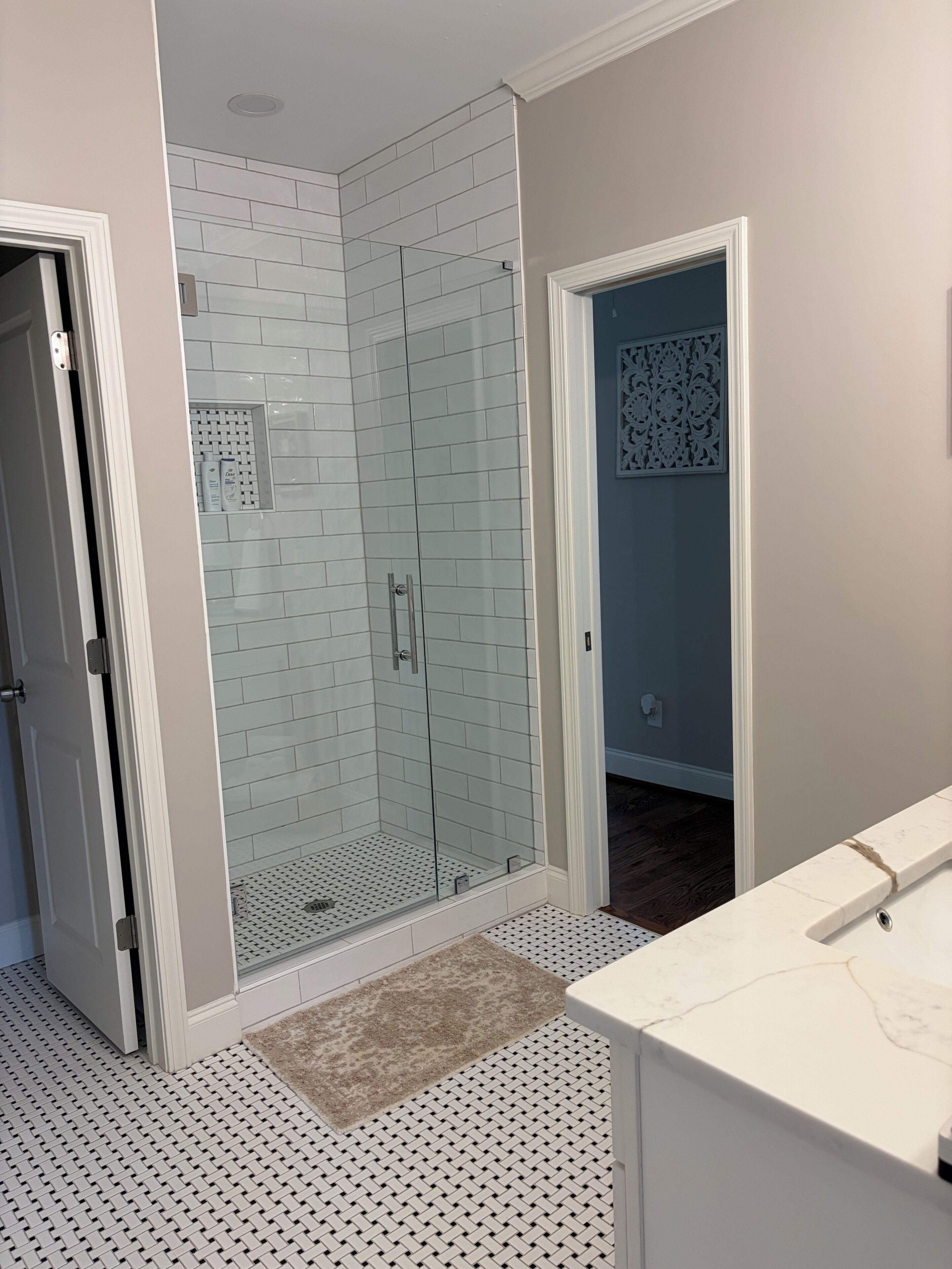 A bathroom with a glass shower enclosure, white tiled walls, a white vanity, and patterned flooring.
