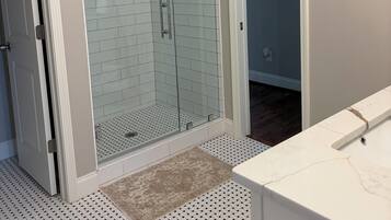 A bathroom with a glass shower enclosure, white tiled walls, a white vanity, and patterned flooring.
