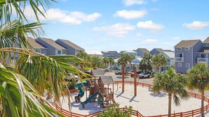 A playground with a slide and swings, surrounded by palm trees and a sandy area, with buildings in the background. - Barrier Dunes 130 - 152 Millie's Dream (Port St. Joe)