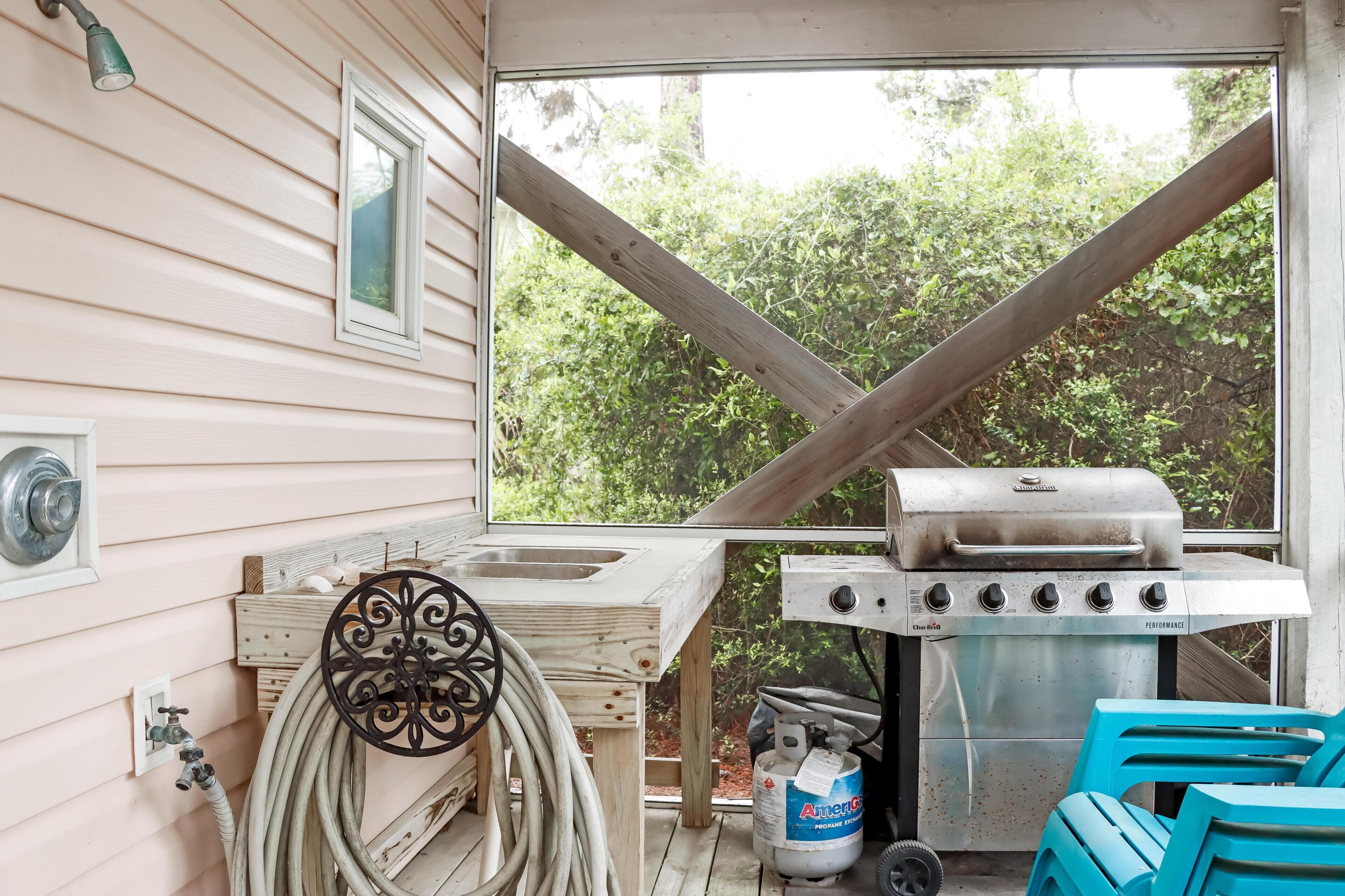 A small outdoor kitchen with a gas grill, a sink, and a window overlooking greenery.