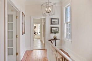 A hallway with a wooden floor, white walls, a geometric chandelier, and framed pictures on the wall. - The Riverview | 1 Block to River St (Savannah)