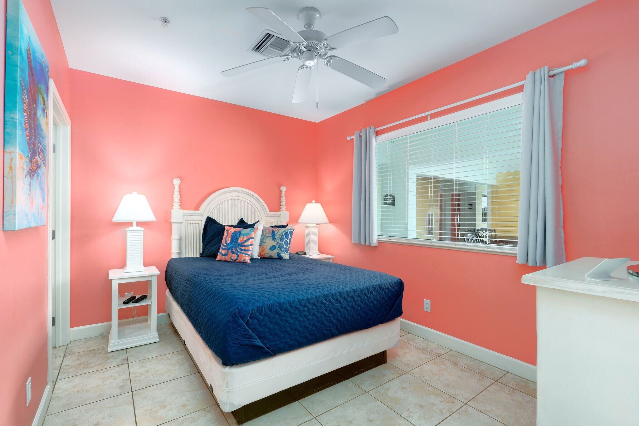 Une chambre à coucher avec un lit, deux tables de chevet avec des lampes, un ventilateur de plafond, une fenêtre avec des rideaux et un tableau au mur.