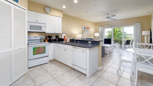 A modern kitchen with white cabinets, a microwave, an oven, and a dishwasher. There is a dining area with a table and chairs, and a balcony with a view of palm trees. - Harbour House #305 "Island Time Harbour" (Fort Myers Beach)