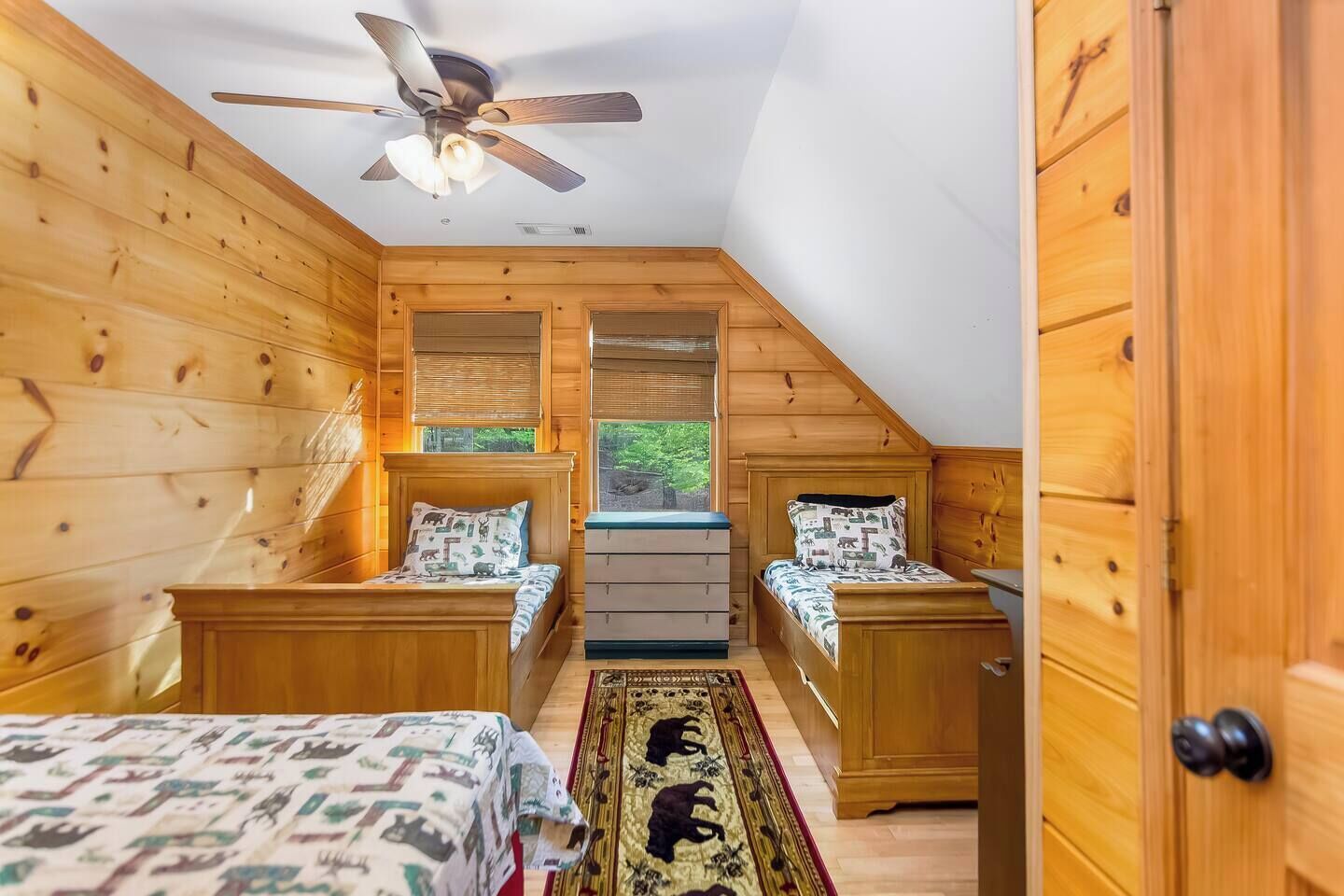 Acogedor dormitorio en el ático con paredes de madera, ventilador de techo, dos camas con ropa de cama estampada, cómoda y vistas a zonas verdes.