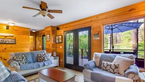 Interior de uma cabana com dois sofás, um ventilador de teto e uma janela grande com vista para o exterior.