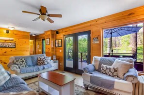 A cabin interior with two sofas, a ceiling fan, and a large window with a view of the outdoors. - Riverfront Escape - Hot Tub • Game Room • Fire Pit (Ellijay)