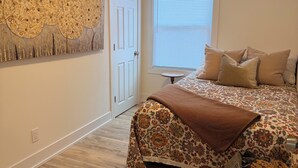 Un dormitorio con suelo de madera, una cama con ropa de cama estampada, un adorno en la pared, una ventana con persianas y una puerta.