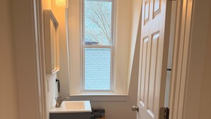 Un pasillo estrecho con una ventana, un lavabo, una cesta y una puerta.