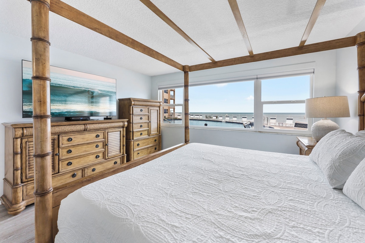 A bedroom with a four-poster bed, a large wooden dresser, a television, and a view of the ocean through large windows.