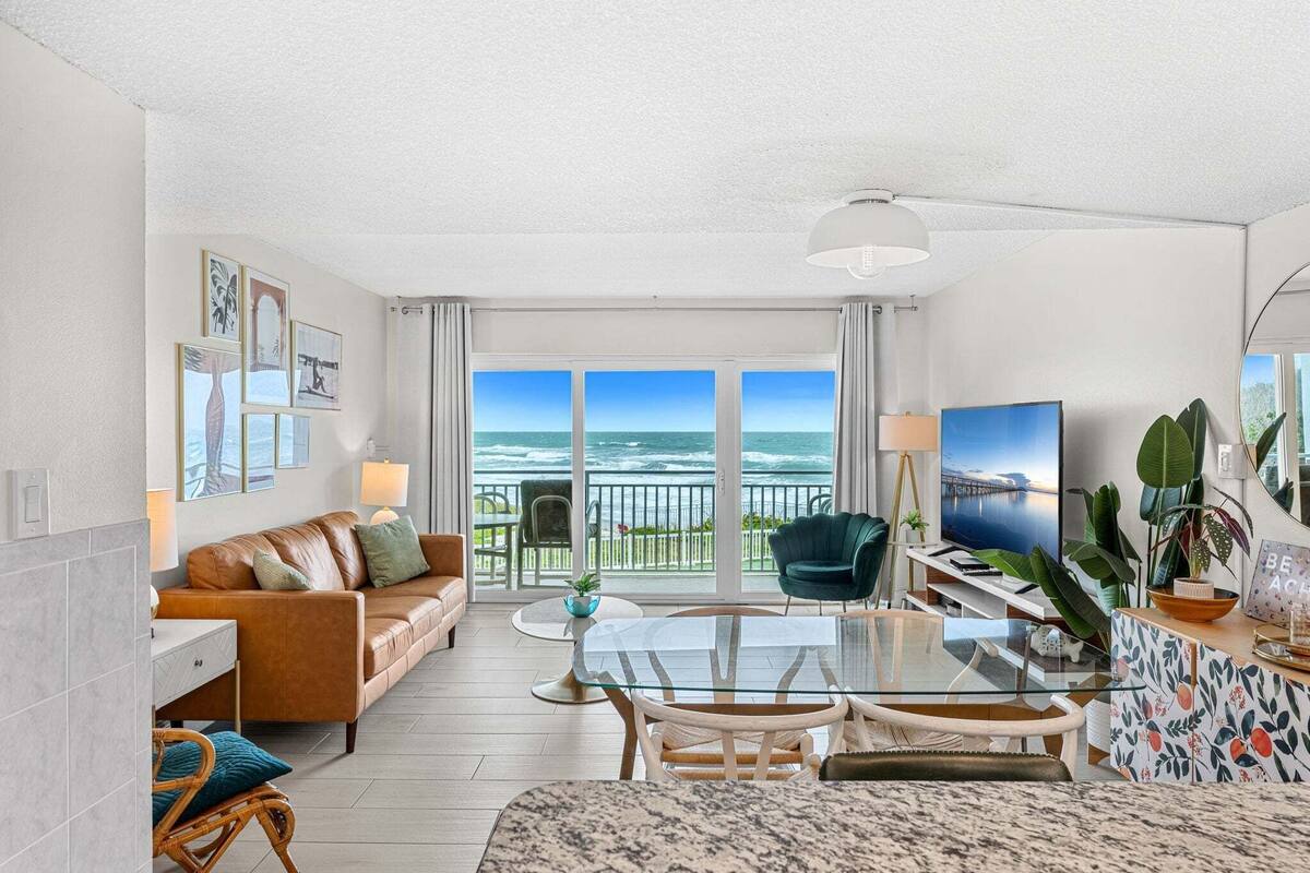 A modern living room with a large window overlooking the ocean, a glass dining table, and comfortable seating.