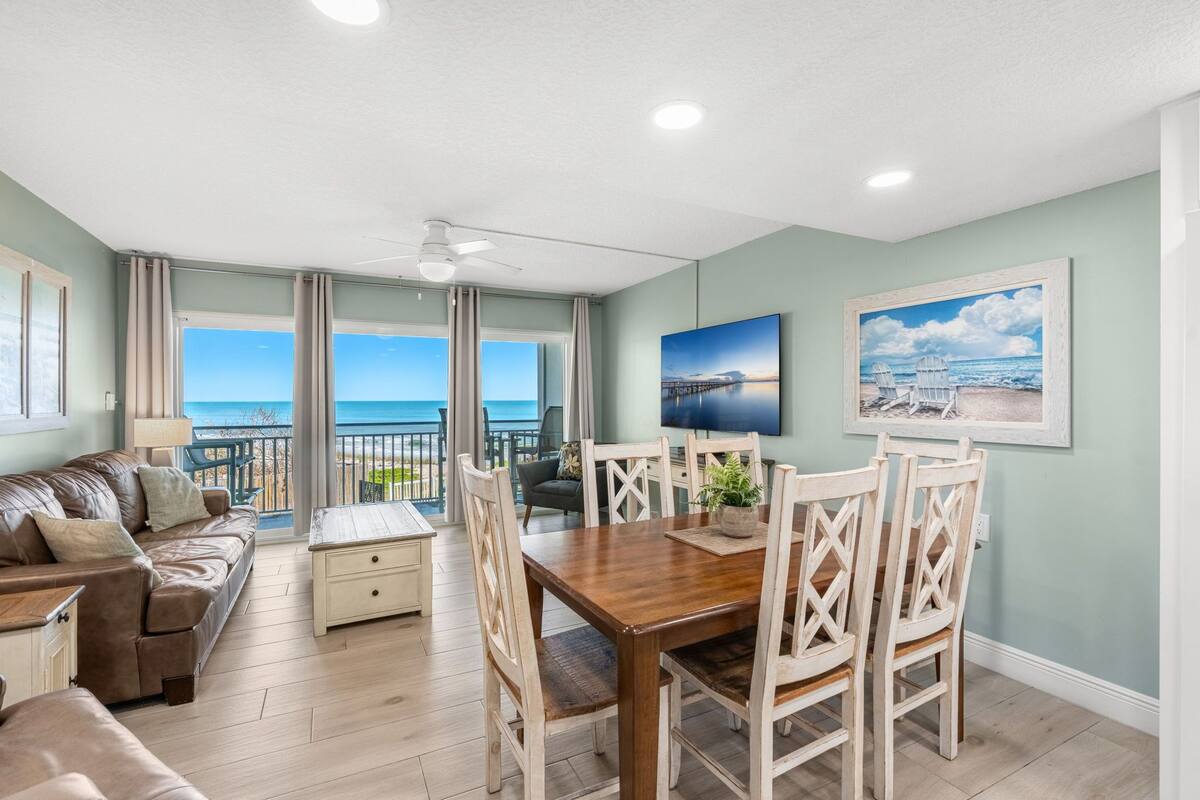 A living room with a dining area, a ceiling fan, a large window with a view of the ocean, and a painting on the wall.