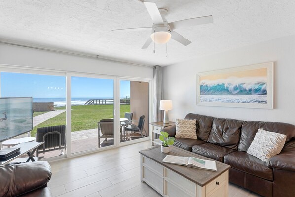 A living room with a large sliding glass door leading to a patio, a ceiling fan, a brown leather couch, a coffee table with a book and a plant, and a framed picture on the wall. - Paradise Beach Club - Direct Oceanfront Condos with Heated Pool Pet-Friendly (Satellite Beach)