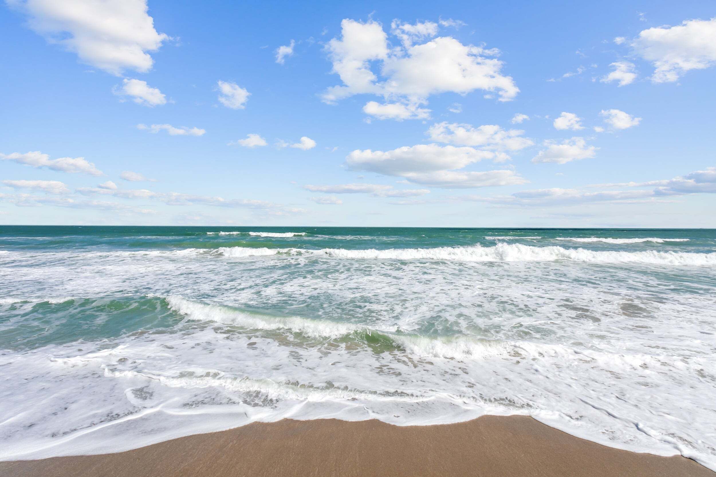 Ein Strand mit Wellen, blauem Himmel und vereinzelten Wolken.