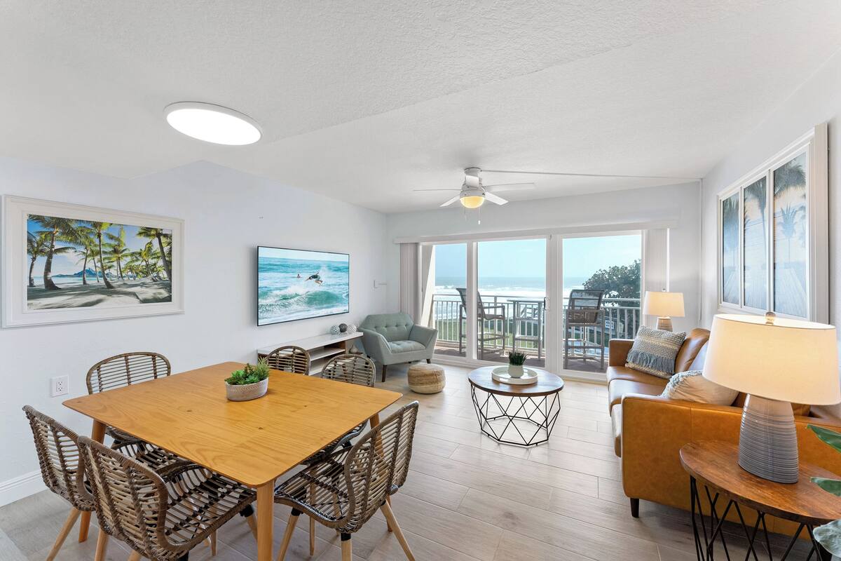 A spacious living area with a wooden dining table, wicker chairs, a grey armchair, a round coffee table, and a sofa with a lamp.