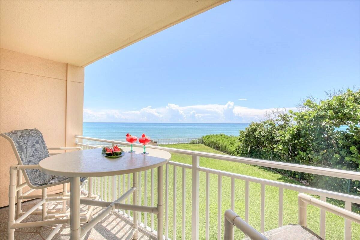 A balcony with a round table and two chairs, overlooking a clear blue sea and a grassy area.