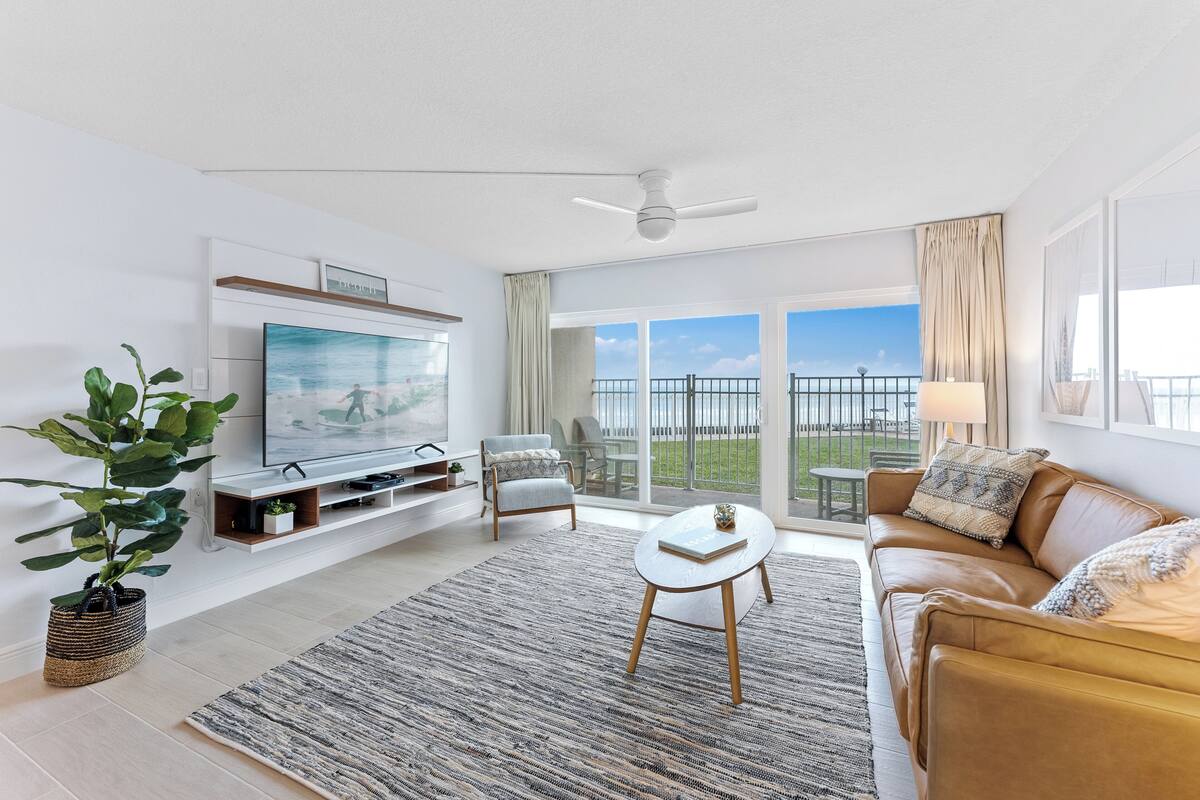 A modern living room with a flat-screen TV, a sofa, a coffee table, and a large window offering a view of the outdoors.